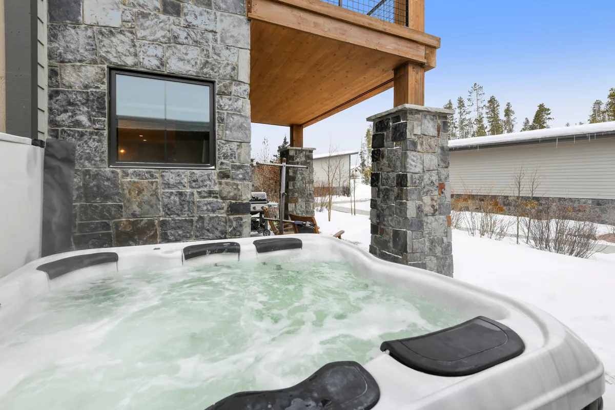 This exterior image showcases a private wooden deck with a prominent hot tub, surrounded by stone pillars. Snow clings to the ground, and the setting conveys a tranquil, luxurious outdoor escape. The scene is inviting, suggesting relaxation and social moments in the hot tub while overlooking the winter mountain scenery.