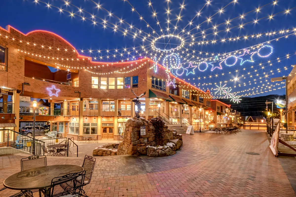 This exterior scene features string lights overhead, decorative stars, an inviting outdoor seating area, and a sculpture fountain, all set within a lively social plaza. The ambiance is joyful and welcoming, suggesting evenings spent dining, socializing, and enjoying community events under festive lighting.