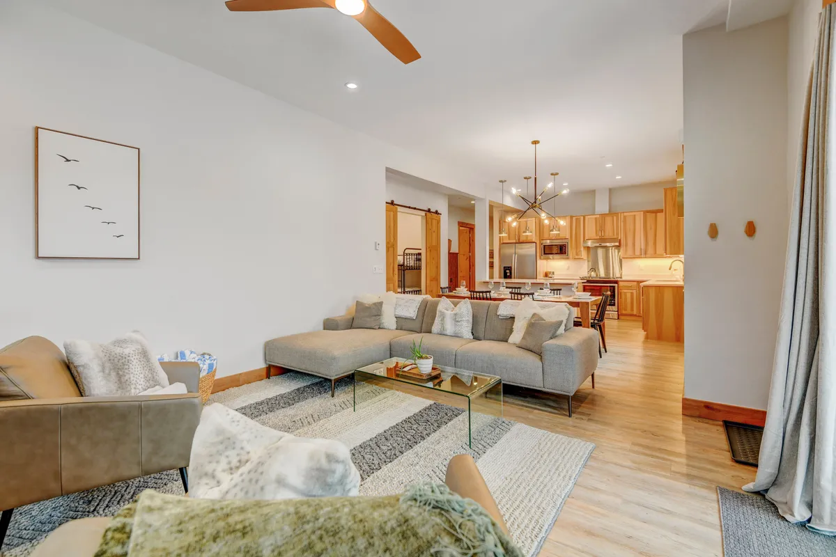This image showcases a large sectional sofa, a stylish coffee table, artwork on the wall, and an open kitchen area in the background. The scene is inviting, featuring a comfortable gathering space with warm, welcoming décor and ample seating for relaxation or socializing.