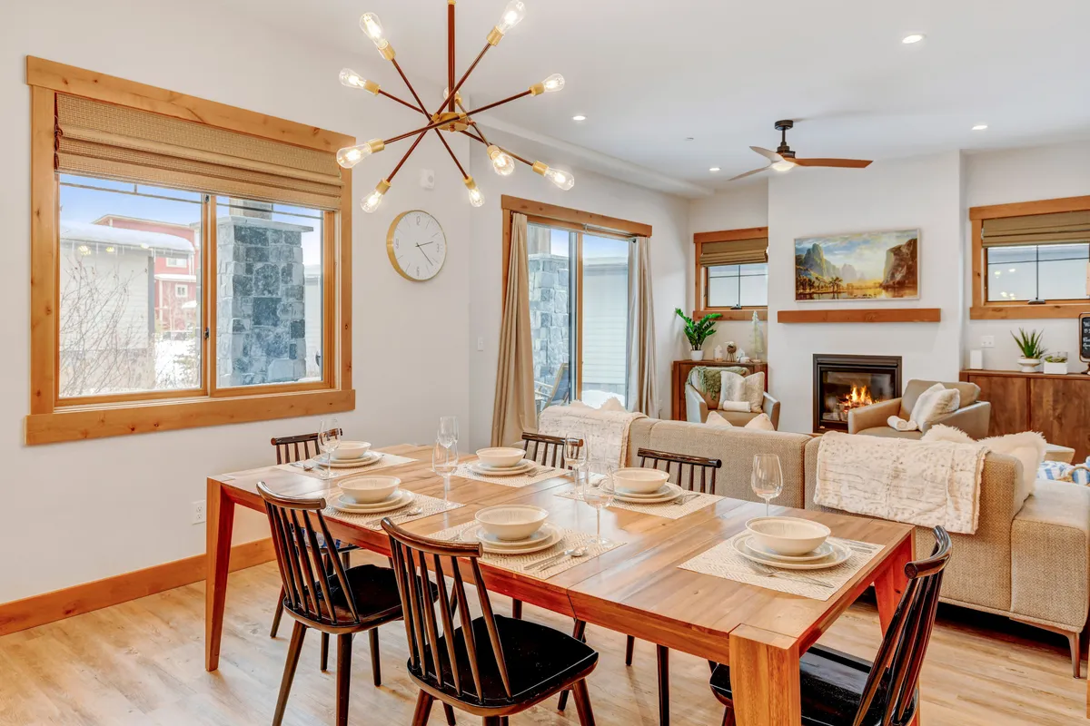 A warm dining area with a central table beneath a chandelier, a fireplace, window views, and a nearby sofa. The space feels inviting and is set up for shared meals, laughter, and relaxation by the fire.