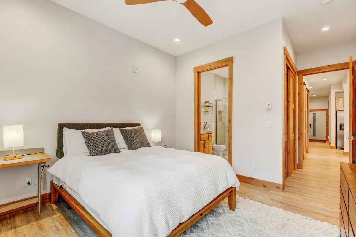 A tranquil bedroom featuring a cozy bed with plush linens, a nightstand, wooden accents, and a doorway that leads directly to an ensuite bathroom. Gentle lighting and soft textures create an inviting, restful atmosphere.