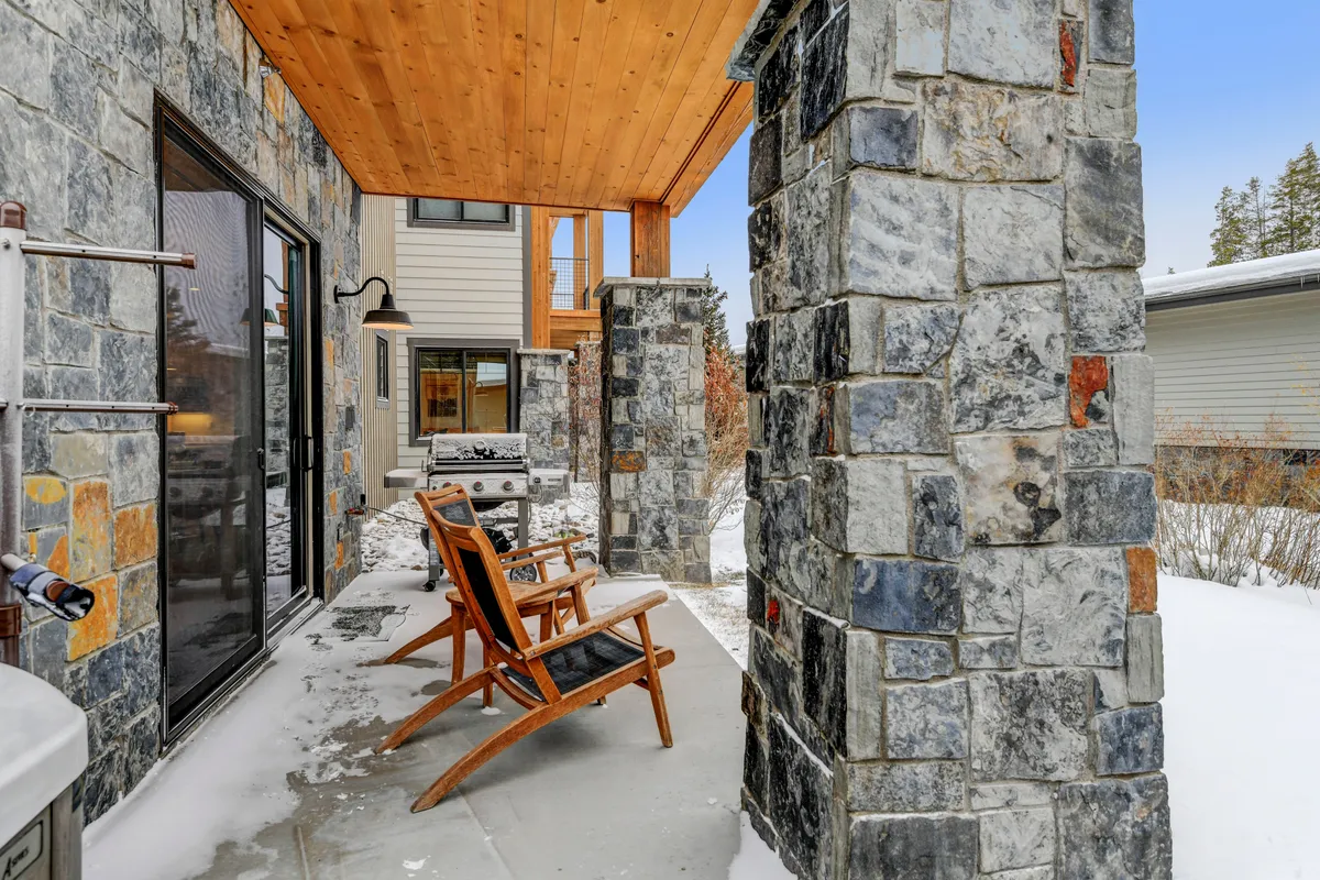A cozy exterior porch is showcased, featuring sturdy stone pillars, a warm wooden ceiling, and inviting rocking chairs. The ground is dusted with snow, suggesting a year-round appeal and the potential for both winter and summer enjoyment. There’s a sense of relaxation and social gathering, with space for enjoying hot drinks, fresh air, or grilling outdoors in a scenic setting.