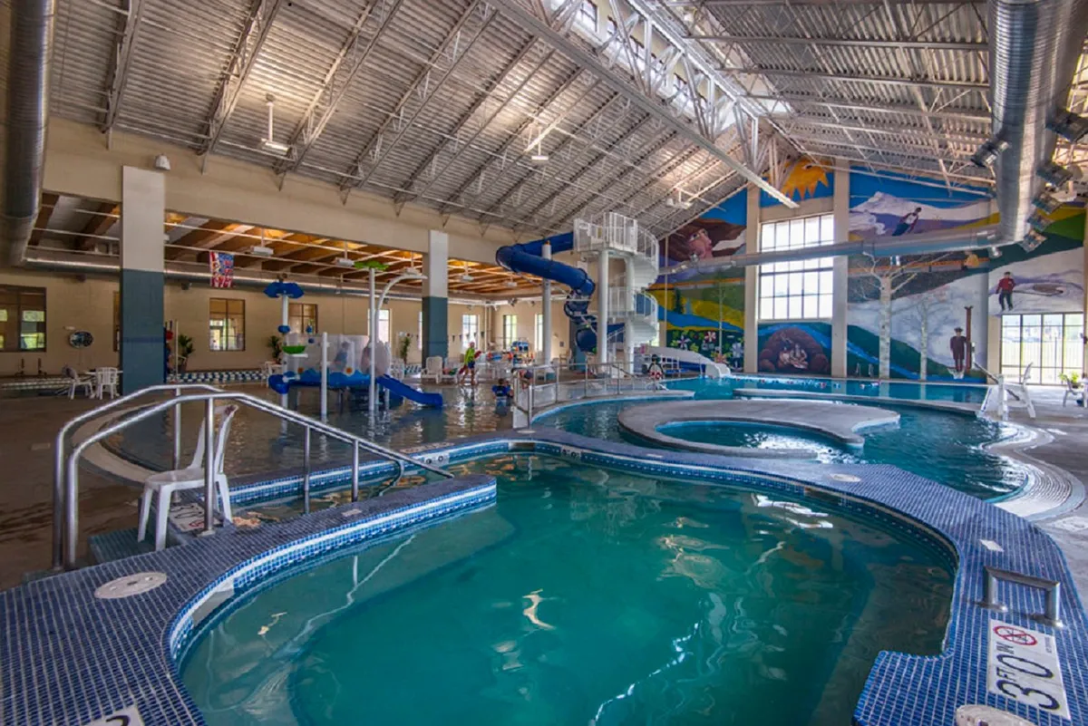 This energetic interior image shows a colorful recreation center with water slides, a swimming pool, and a vibrant mural, bustling with kids and families enjoying aquatic fun. The scene suggests a joyful, kid-friendly environment perfect for year-round recreation.