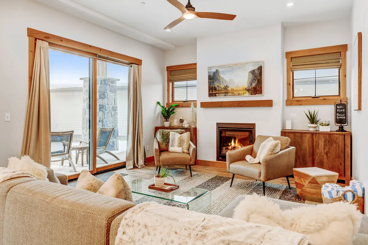 The image showcases a cozy, beautifully decorated living room with a glowing fireplace as the focal point, a plush sofa adorned with blankets, tasteful artwork above the mantle, and a sliding glass door leading to a patio with scenic sunset views. Decorative plants add warmth and charm, creating a space perfect for unwinding after a day of adventure.